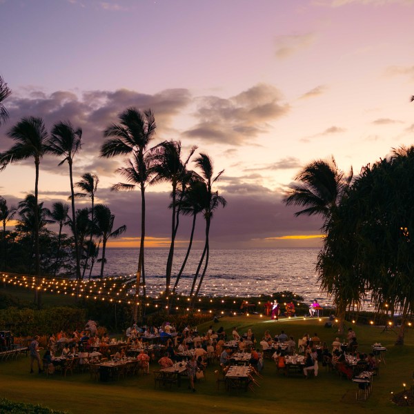 Outdoor event at sunset with palm trees, string lights, and ocean view.
