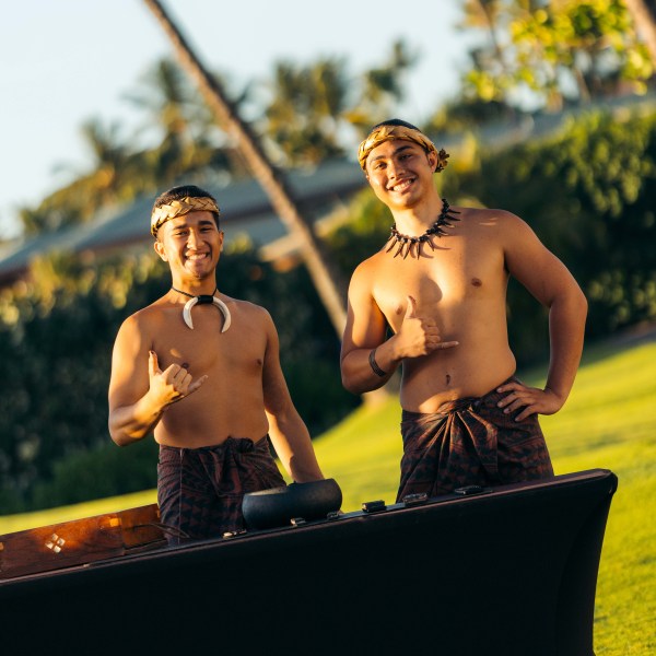 Two men in traditional attire giving shaka sign outdoors with palm trees.