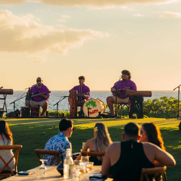Five musicians in purple shirts perform outdoors near the ocean at sunset.