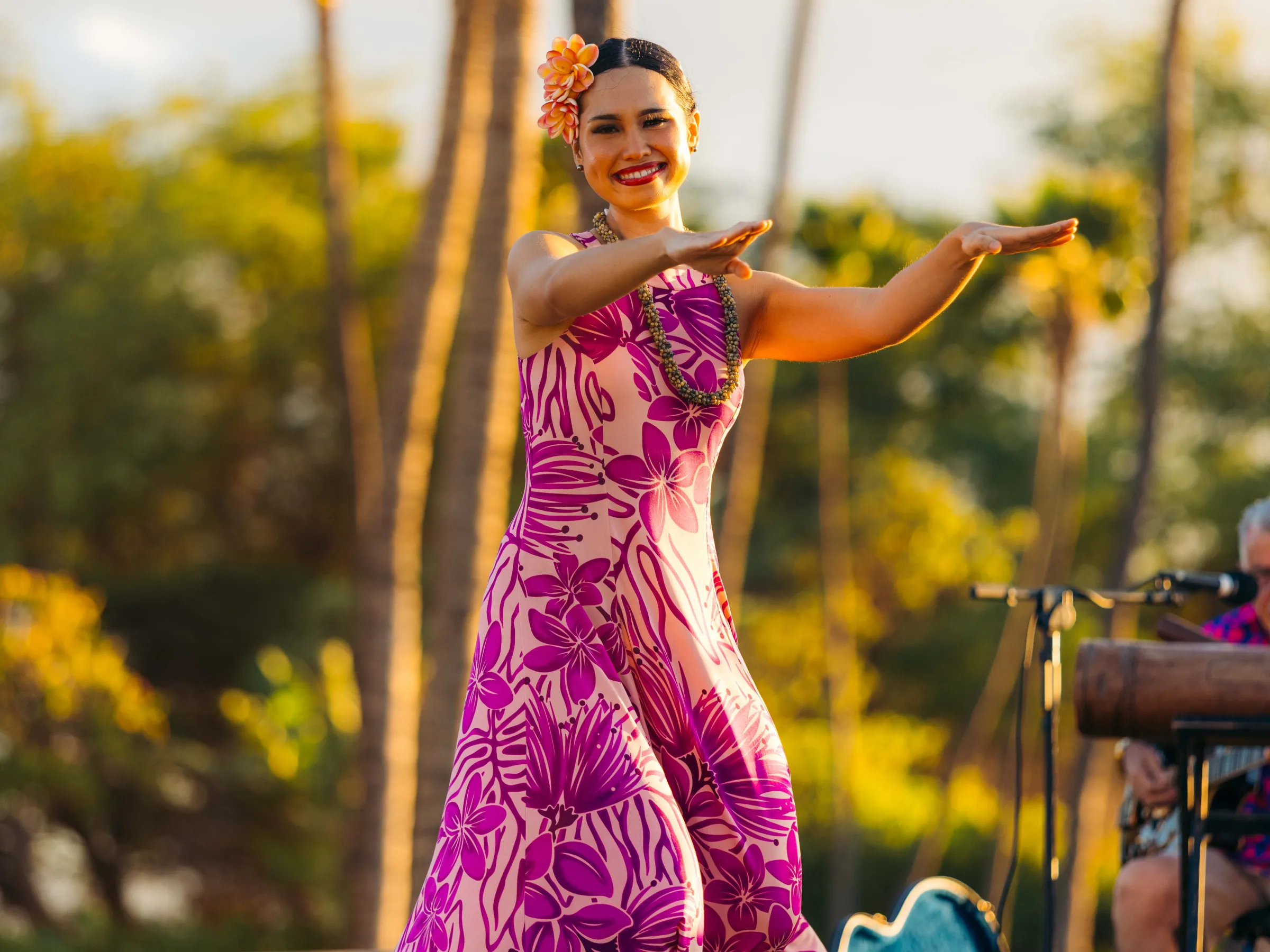 Woman in pink floral dress dancing hula outdoors.