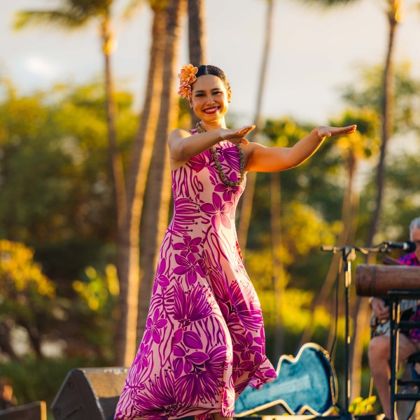 Woman in pink floral dress dancing hula outdoors.