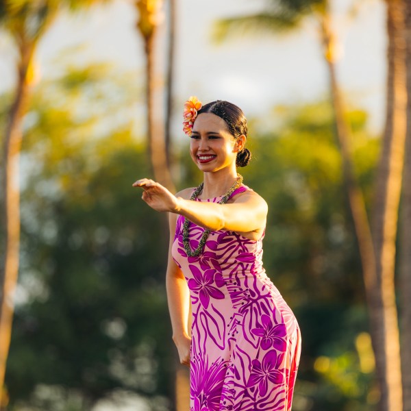 Woman in pink floral dress dancing hula outdoors, palm trees in background.