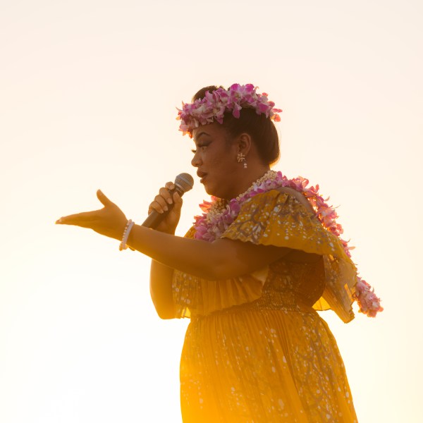 Person in floral dress and lei speaking into a microphone at sunset.