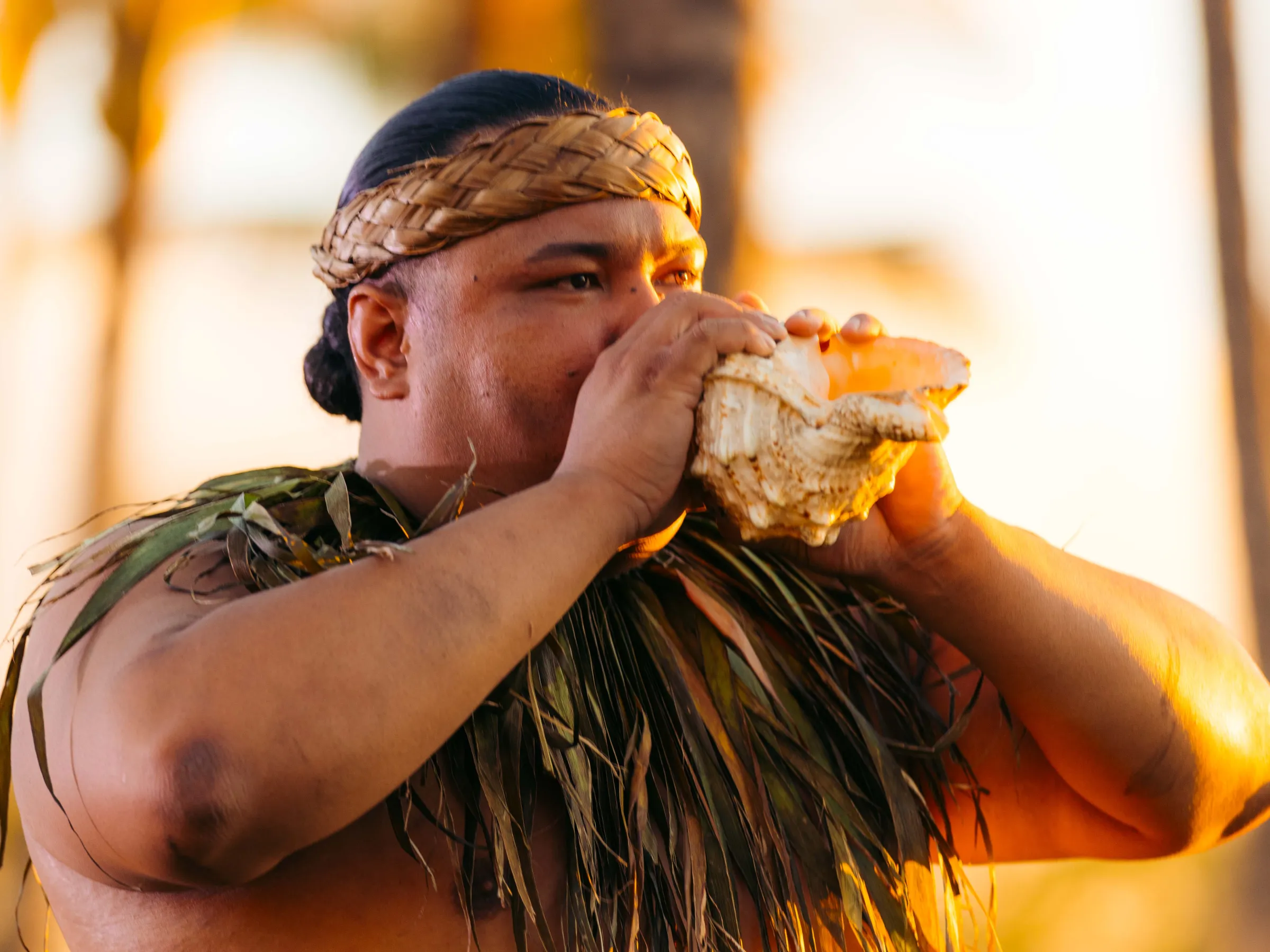 Person blowing a conch shell, wearing traditional attire with palm trees in the background.