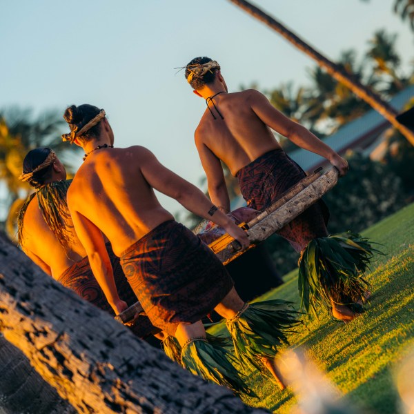 Three people in traditional attire carrying a log outdoors, with palm trees in the background.