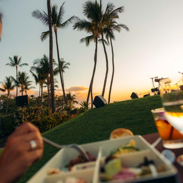 Person eating a meal outdoors with palm trees and sunset in the background.