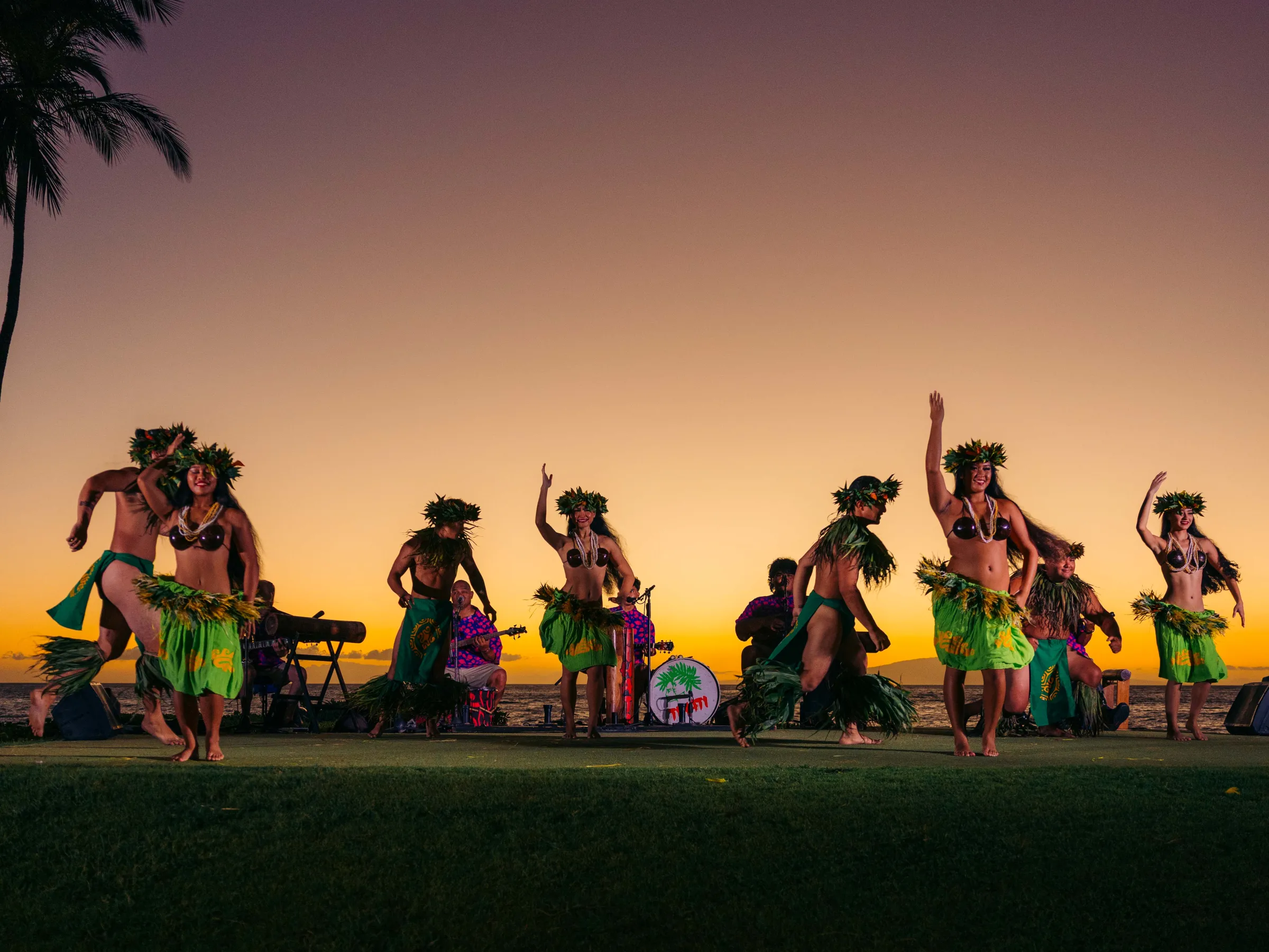 Hula dancers perform at sunset with a band in the background.