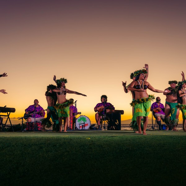 Hula dancers perform in grass skirts with musicians at sunset.