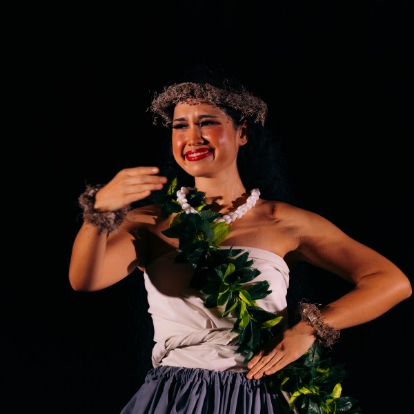 Dancer in traditional attire with lei against dark background.