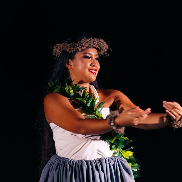 Woman in traditional attire performing a hula dance against a dark background.