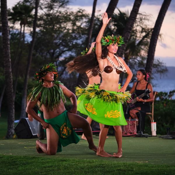 Two dancers in traditional attire perform with musicians outdoors at sunset.