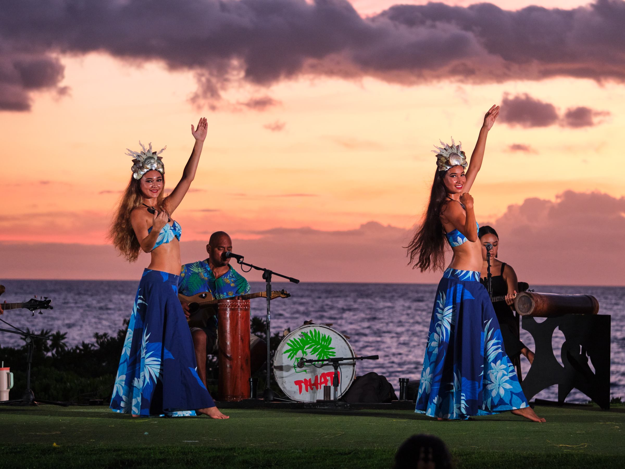 Two women in blue dresses dancing by the ocean at sunset, with musicians playing in the background.