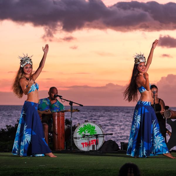 Two women in blue dresses dancing by the ocean at sunset, with musicians playing in the background.