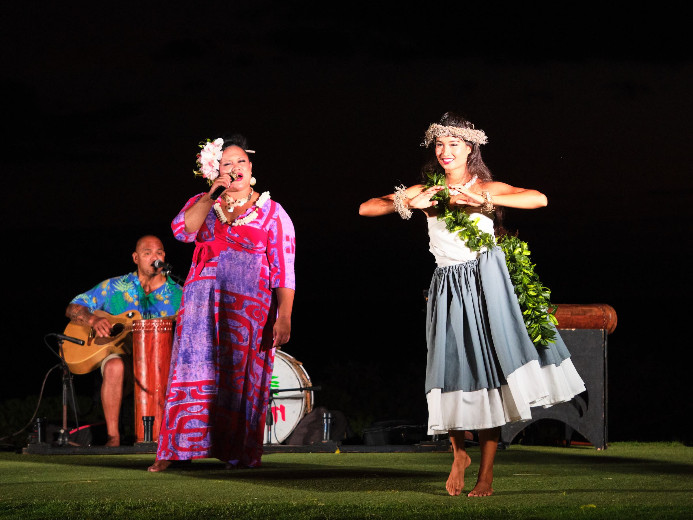 Three performers at night: a dancer in grass skirt, a singer, and a guitarist.