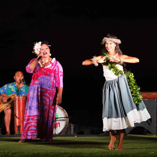 Three performers at night: a dancer in grass skirt, a singer, and a guitarist.