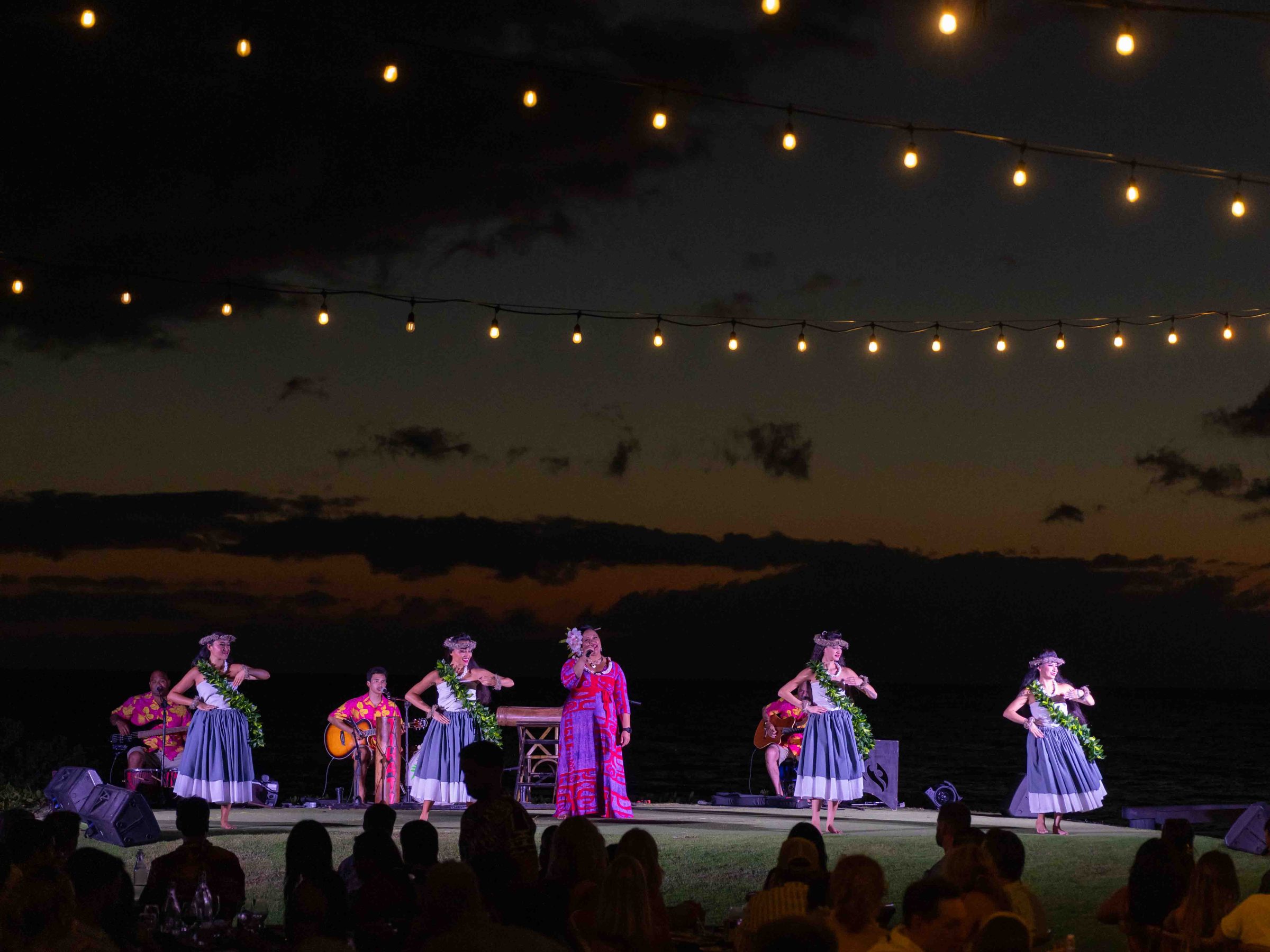 Dancers perform at night under string lights with an audience watching.